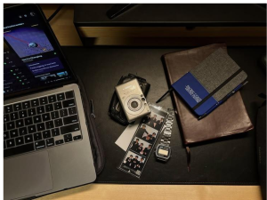 Foto dari atas ke bawah yang melihati laptop, kamera, jam tangan, strip foto, dan buku catatan di atas meja. Biaya barang-barang tersebut akan terpengaruhi oleh melemahnya nilai rupiah.